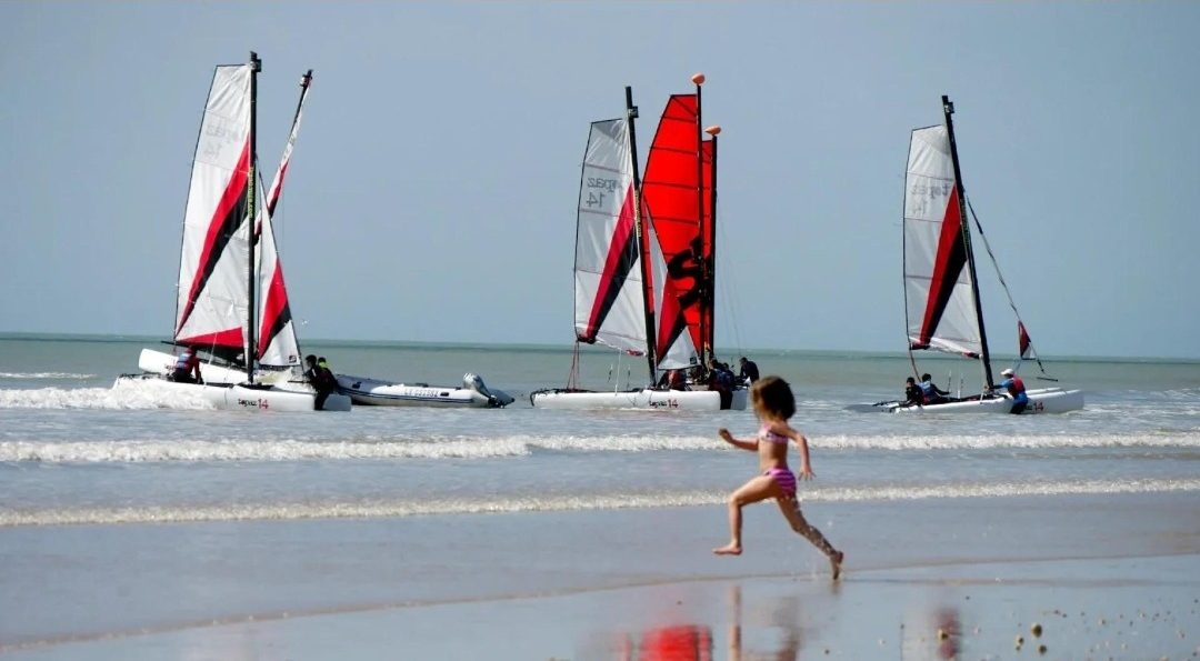 Catamarans aux voiles rouges et blanches au bord de l’eau, une enfant court sur le sable