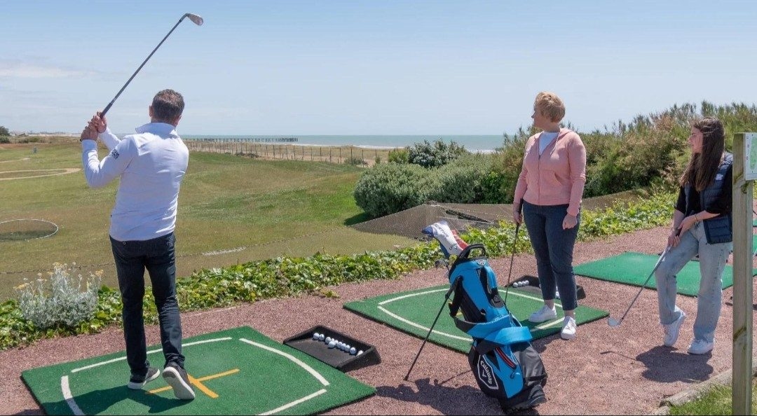 Un golfeur au practice face à la mer, deux personnes le regardent