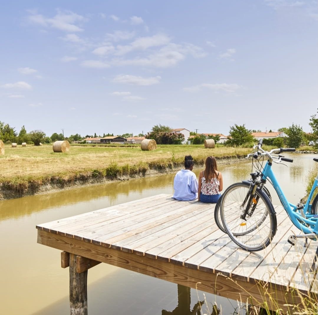 Deux personnes assises sur un ponton au bord d’un canal du marais, un vélo à côté